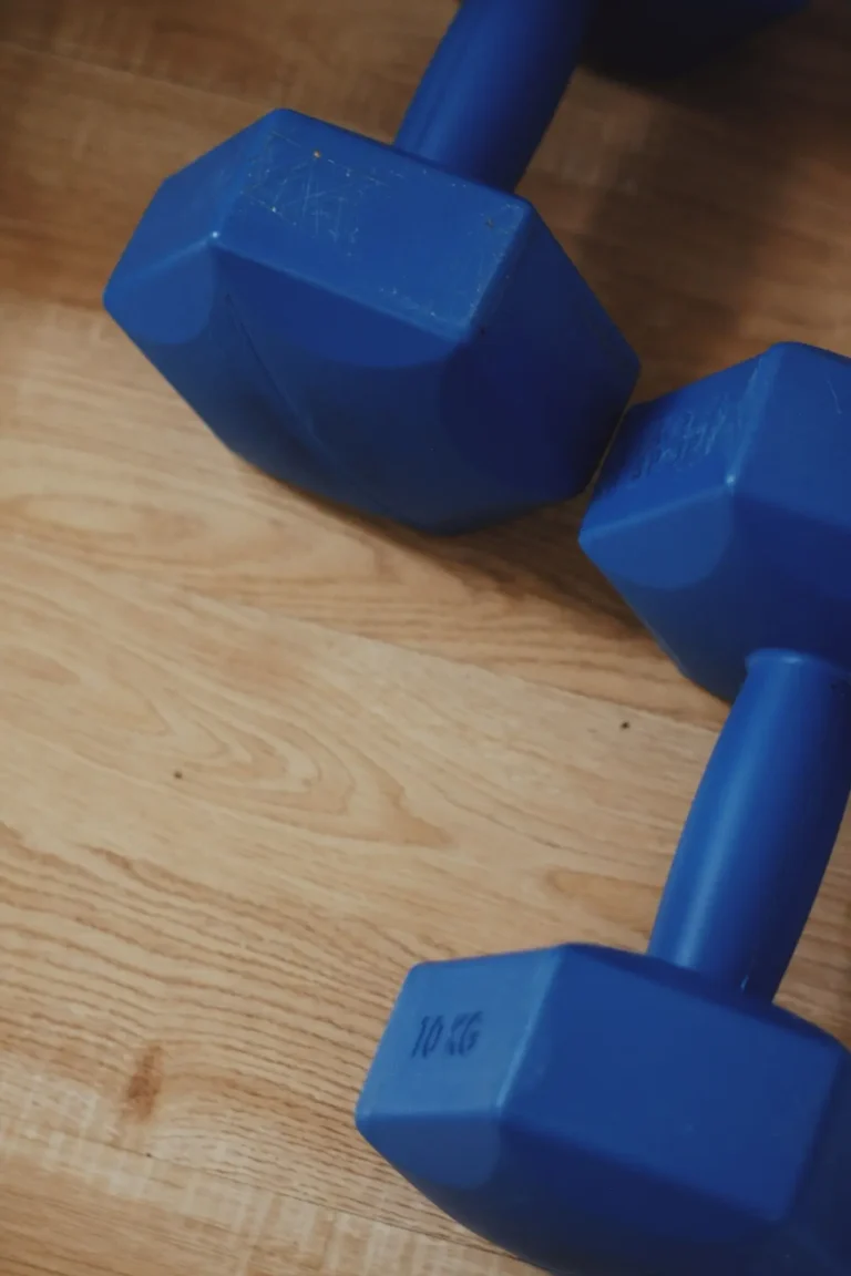 Two blue hexagonal dumbbells resting on a wooden floor, each marked “10 KG,” representing simple strength‑training equipment used in exercise.
