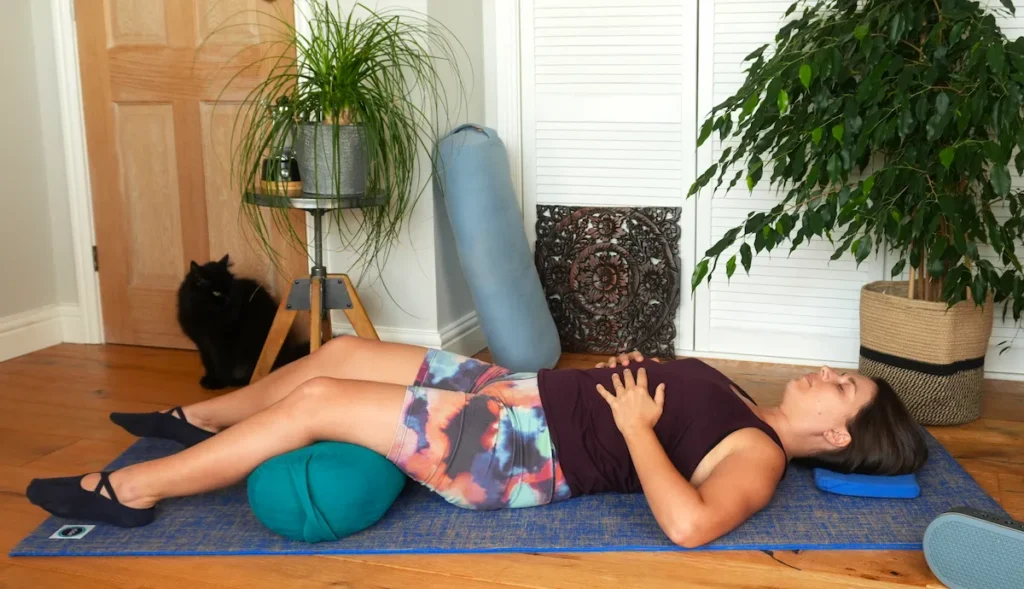 Person lying in a supported relaxation pose on a yoga mat with cushions and a bolster, in a calm indoor space with plants and a cat nearby.