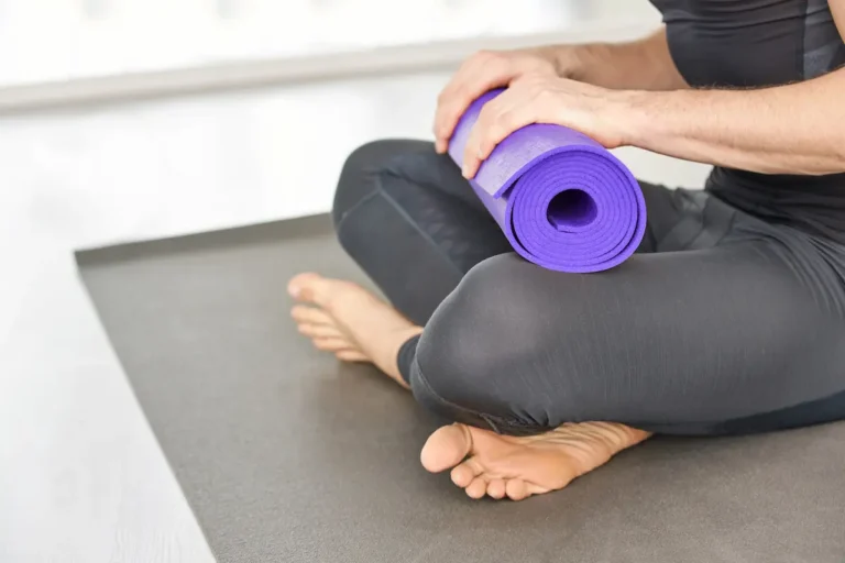Person sitting cross‑legged on a yoga mat holding a rolled mat, preparing for exercise.
